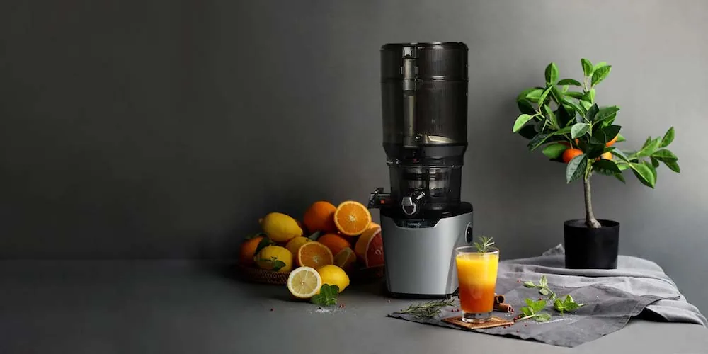 Kuvings AUTO10S Plus Slow Juicer aufgebaut zwischen Pflanzen, Obst und fertigen Säften